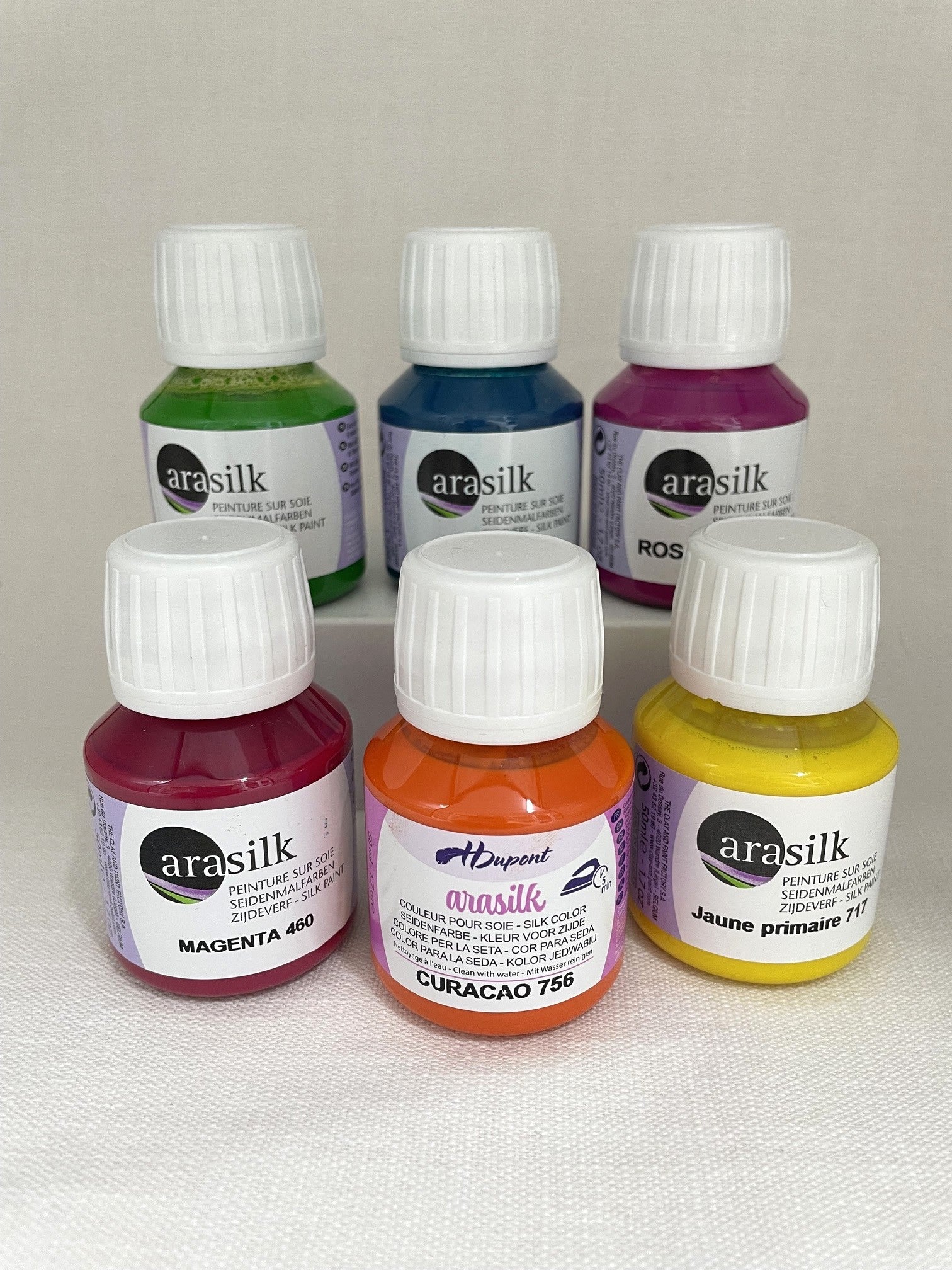 Six small bottles of Arasilik silk paint in primary colors, arranged in two rows on a light fabric background. The bottles contain green, blue, purple, magenta, orange, and yellow paint, with white ribbed caps and white labels.