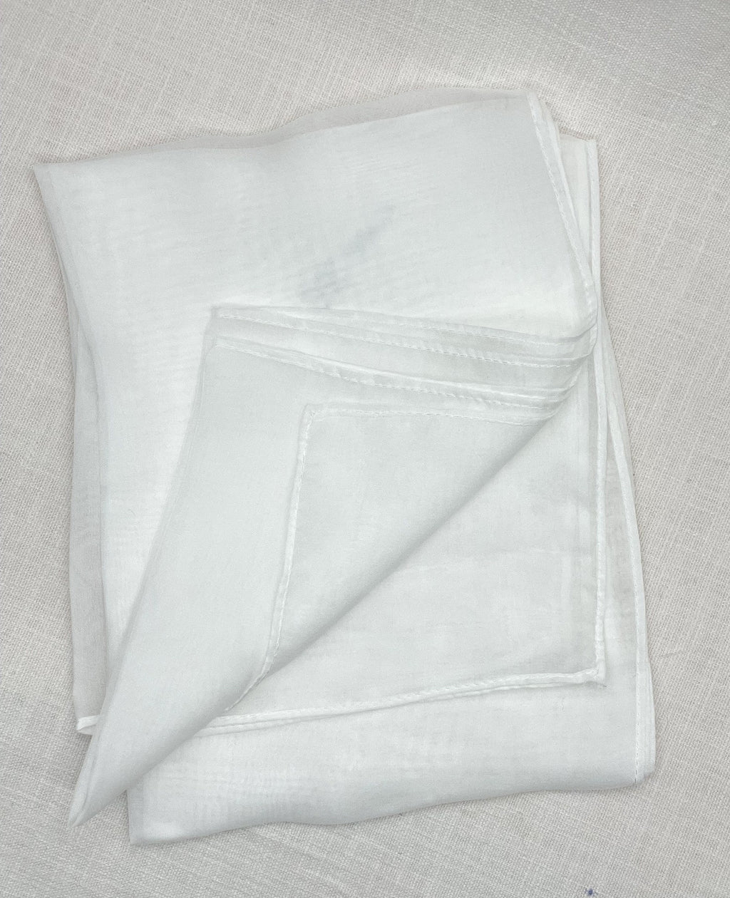 A folded white chiffon scarf with a stitched border, displayed on a textured beige linen surface.