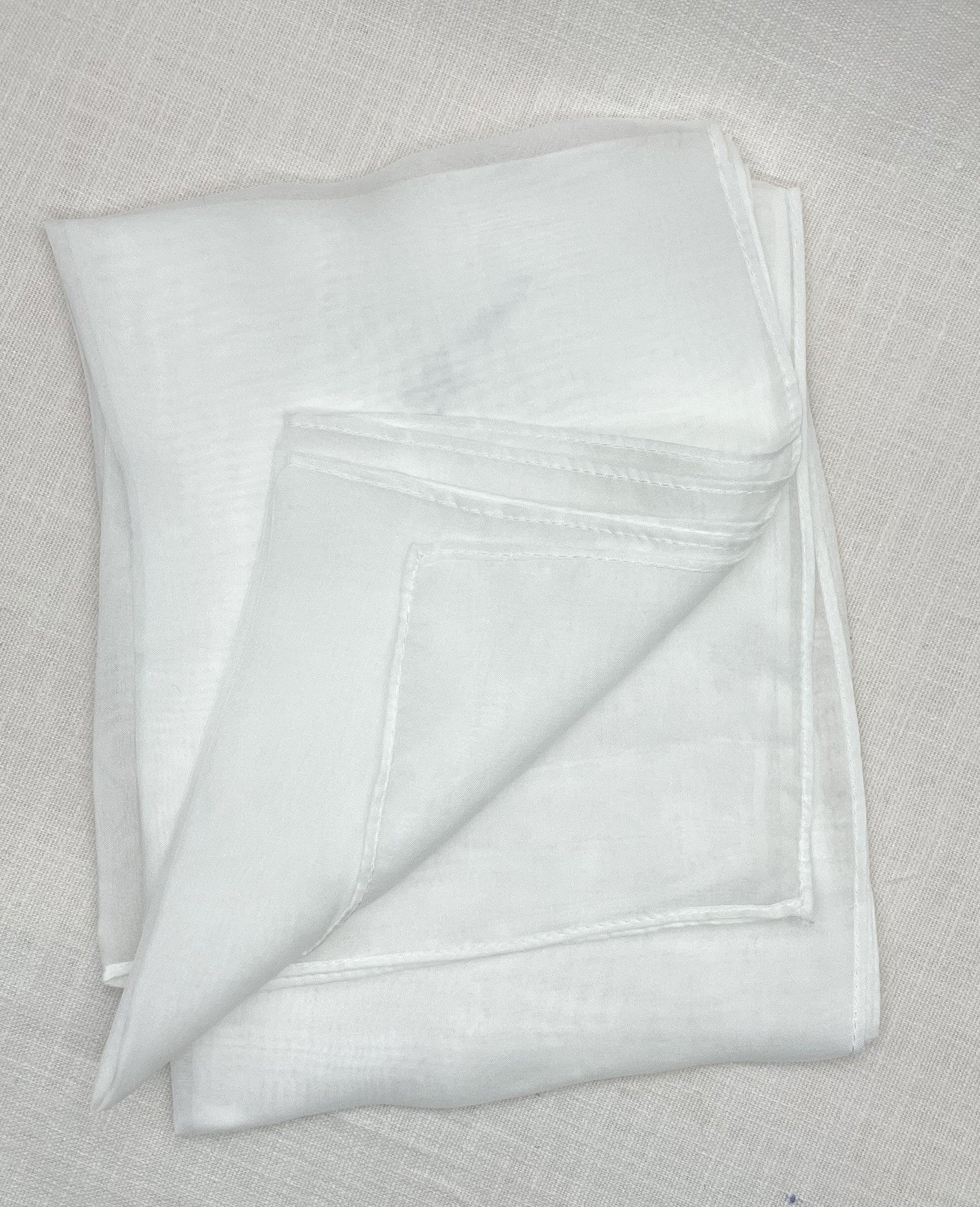 A folded white chiffon scarf with a stitched border, displayed on a textured beige linen surface.