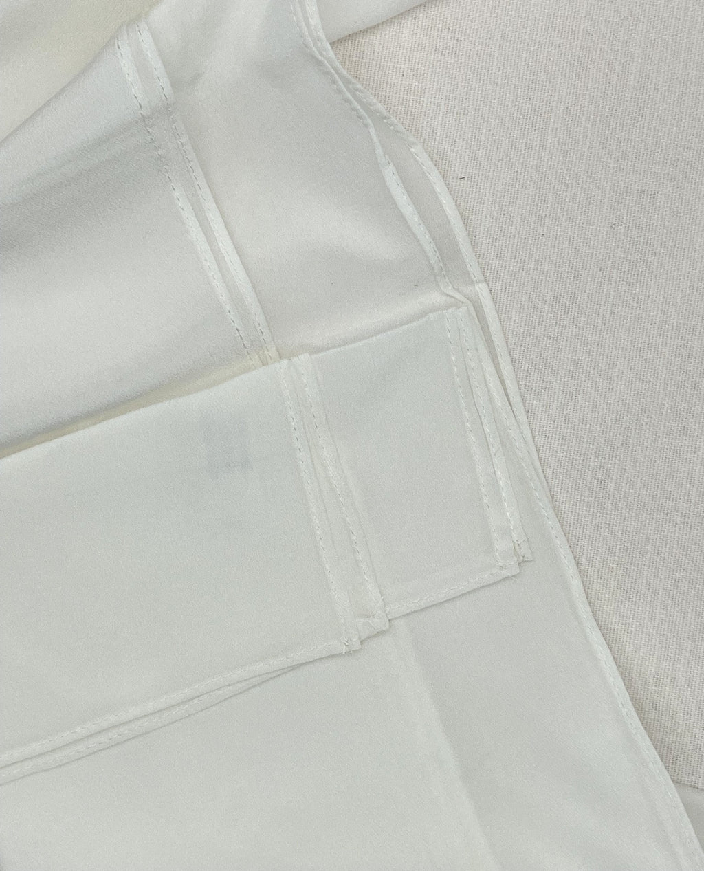 Close-up of a undyed machine hem scarf with visible seams and a textured off-white woven fabric background.
