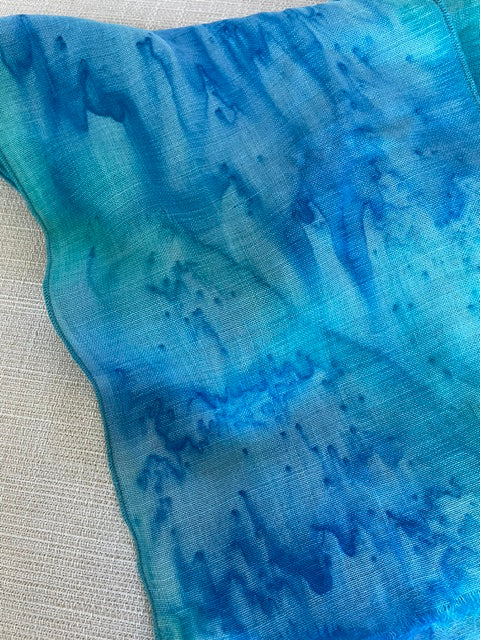 A close-up of a lightweight wool gauze scarf dyed blue by Ditchfield Designs with a watercolor or tie-dye pattern, draped diagonally over a light beige textured surface.
