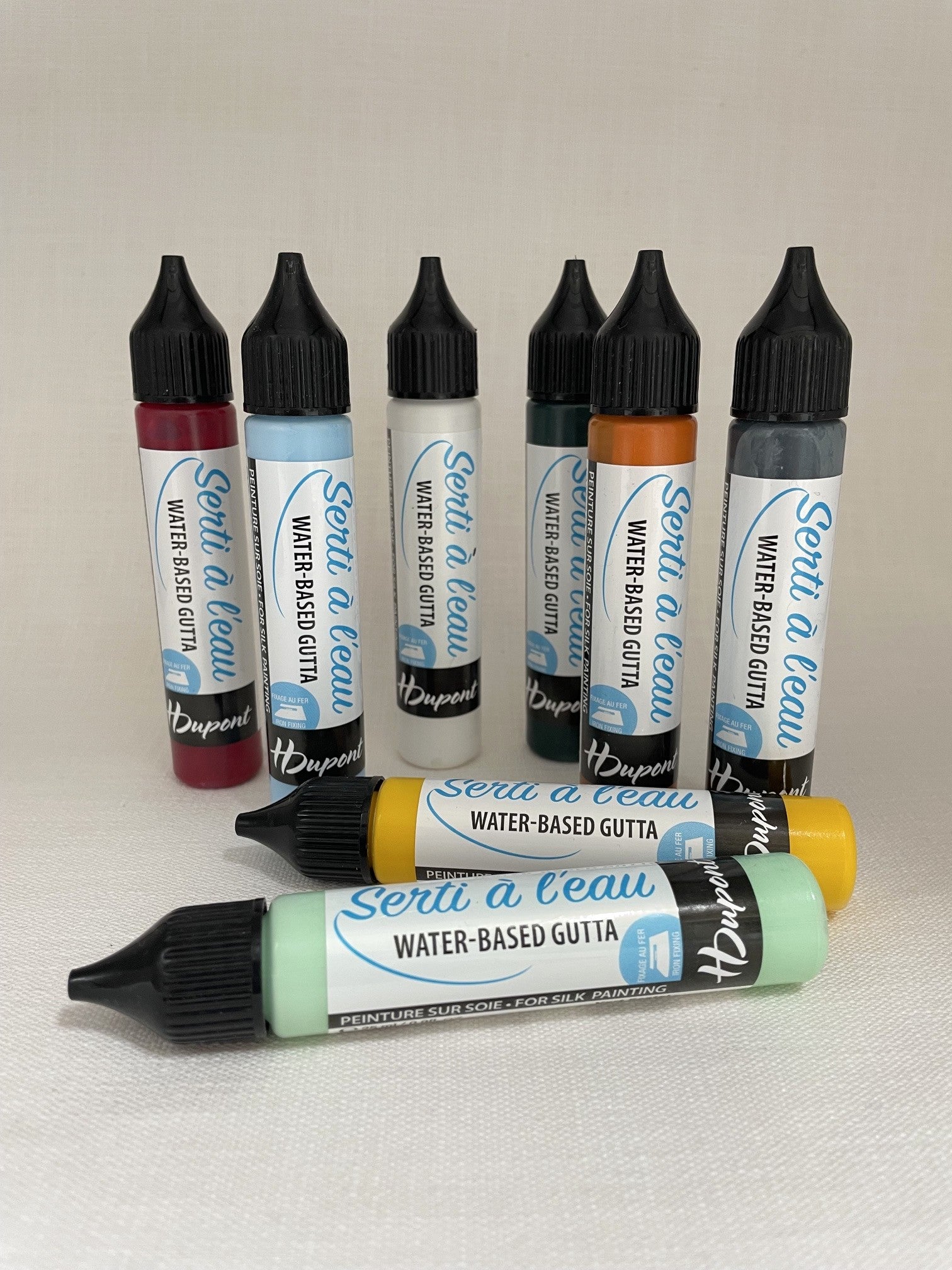 Six bottles of water-based gutta arranged on a light beige fabric background. The set includes red, light blue, white, dark teal, orange, and gray bottles, with two bottles in the foreground showing yellow and mint green caps.