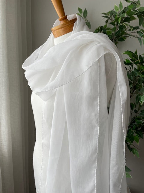 Undyed white crinkle chiffon silk scarf on a wooden hanger with green plants in the background