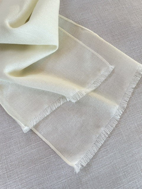 An undyed lightweight wool scarf with fringed edges, draped elegantly on a textured light gray linen surface.