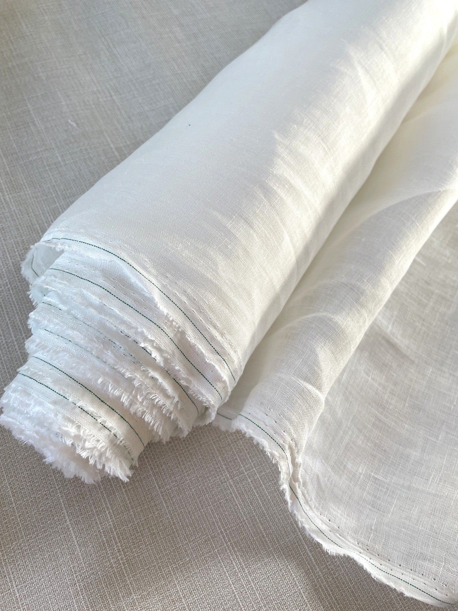 A close-up of a rolled piece of undyed lightweight linen fabric with a raw, frayed edge, laid on a textured light beige linen surface.