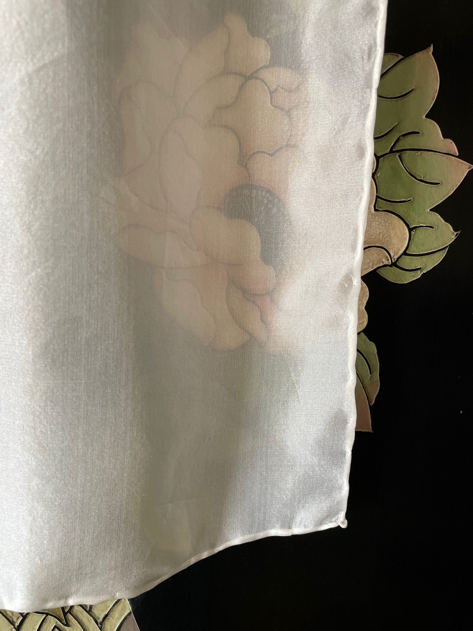 Undyed white Paj silk scarf with a floral design partially visible underneath, against a dark background.