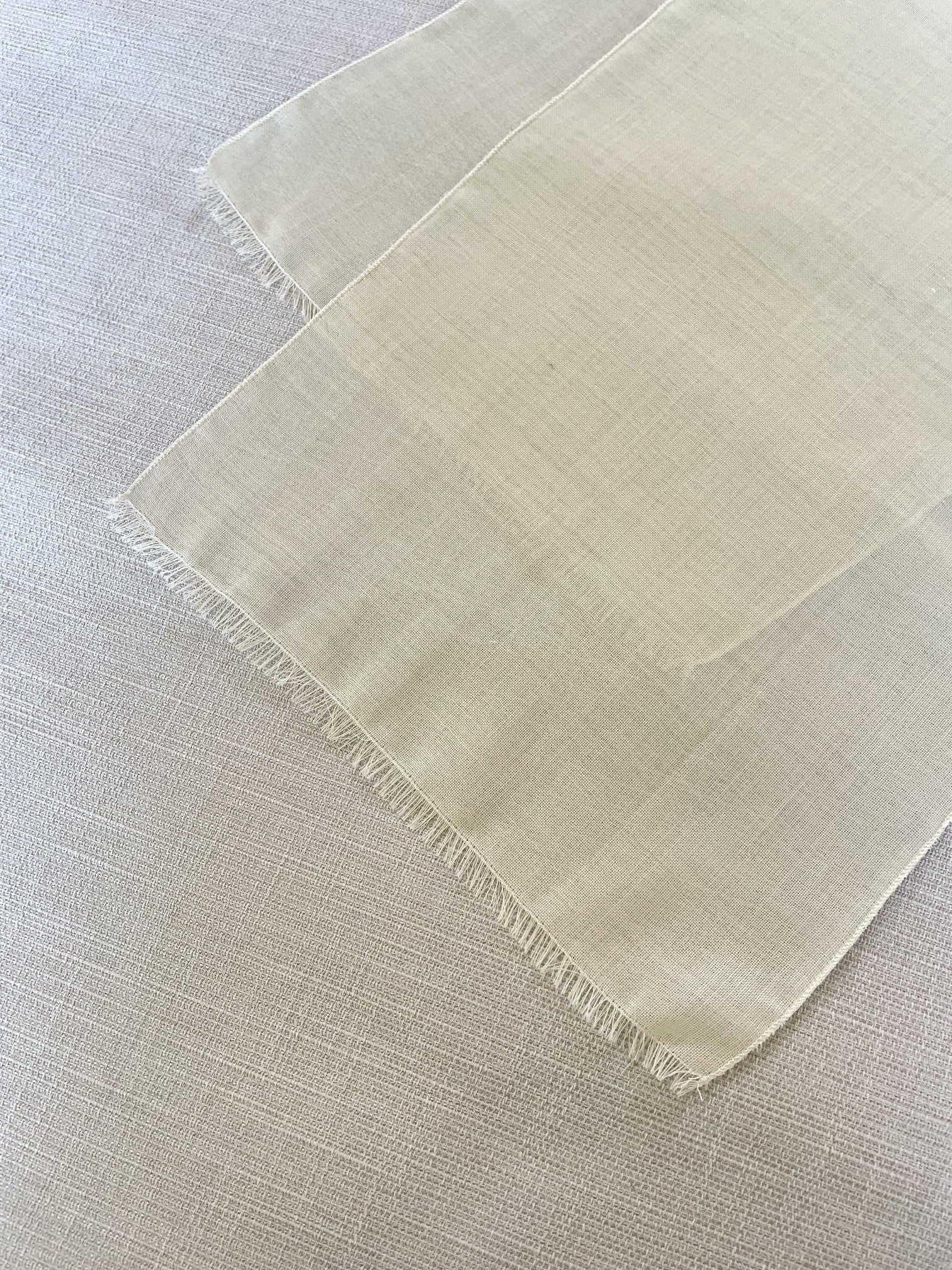 A close-up of a light beige undyed woven wool gauze textile, likely a scarf, with frayed edges, laid flat on a matching textured fabric background.