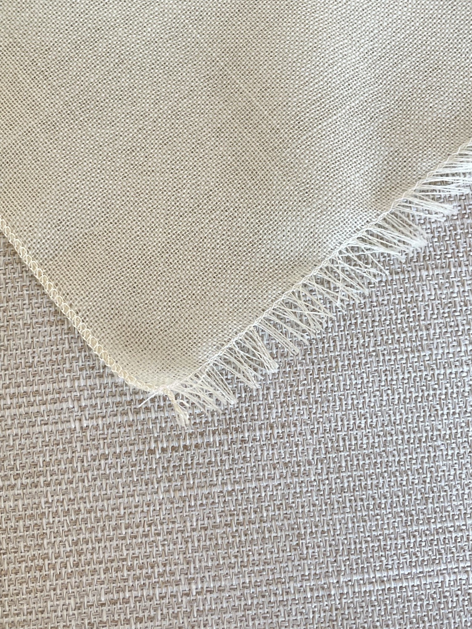 A close-up of a light beige undyed wool gauze woven fabric with fringed edges, laid over a textured neutral fabric background.
