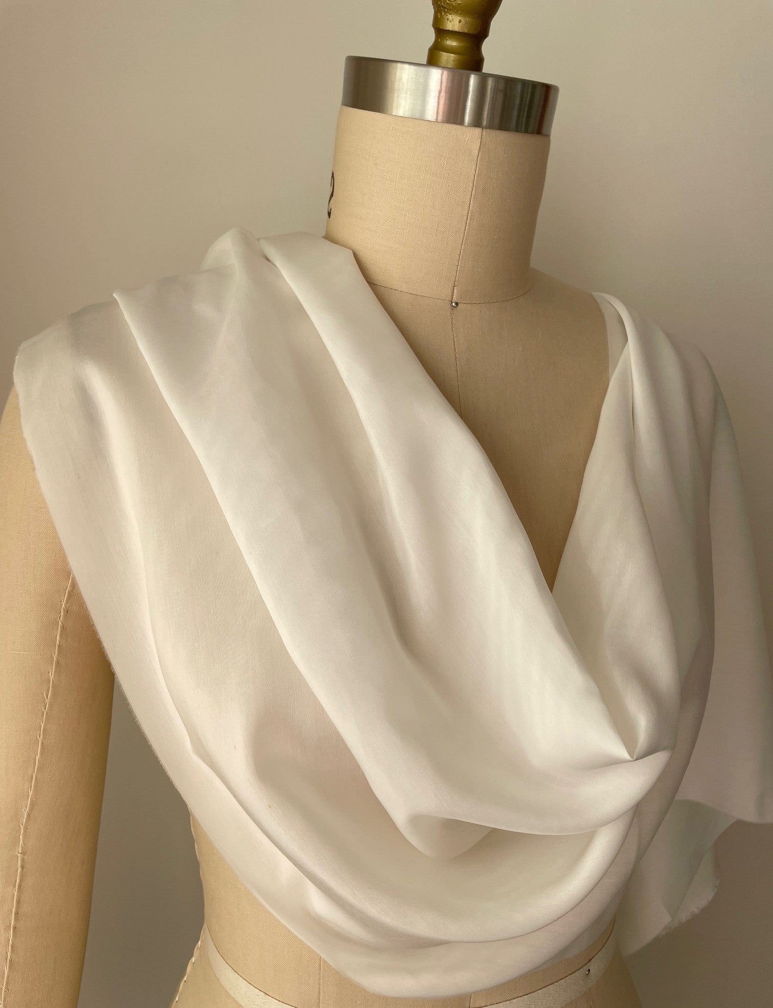 A draped undyed white silk bamboo fabric displayed on a beige fabric dress form mannequin against a plain light beige wall.