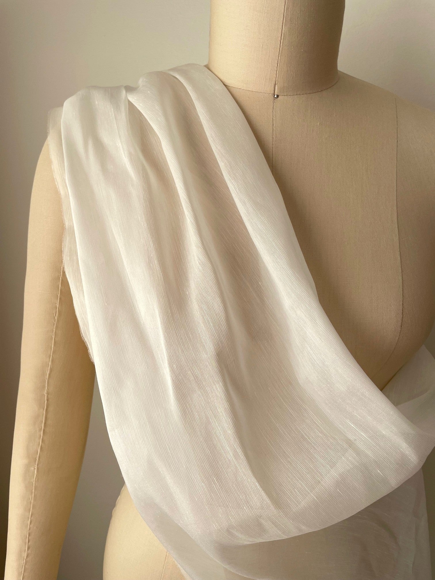 A lightweight, semi-sheer undyed white silk linen fabric draped over a beige mannequin torso, showing its soft texture and flow.