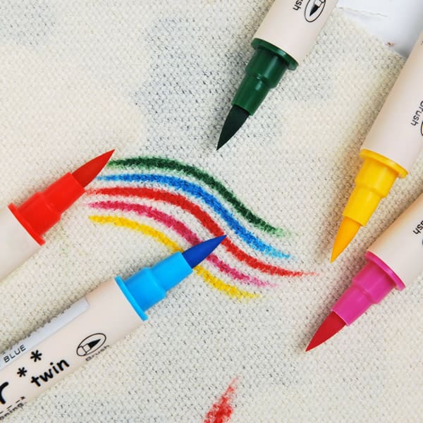 Four twin-tip marker pens in red, blue, green, and yellow are arranged on a textured off-white fabric, with their tips touching to draw a rainbow arc.